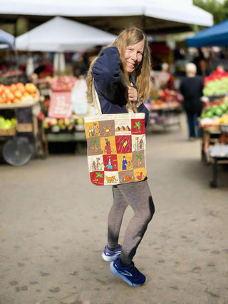Person holding a colorful tote bag with a market scene design at an outdoor market.
