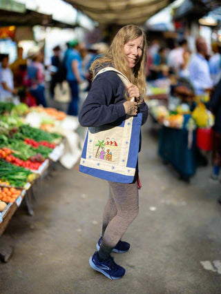 Woman holding a decorated bag in a market setting with vegetables and people in the background