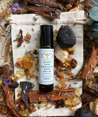 A roll-on essential oil bottle labeled 'Allspice Oudh Natural Botanical Perfume Essential Oil Blend Roll-On' placed on a beige cloth surrounded by dried herbs, crystals, and bark.