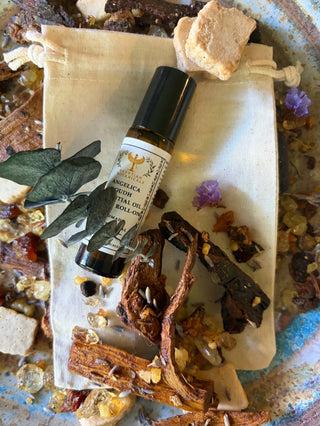 A small amber glass roll-on bottle with a black cap and a cream label reading Angelica Oudh Natural Botanical Perfume styled on a cream drawstring pouch with dried botanicals, bark, flowers, and wax cubes.