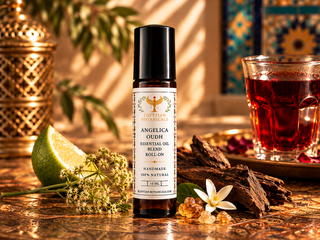 A 10 ml amber roll-on bottle of Angelica Oudh Botanical perfume surrounded by natural ingredients angelica flower,  agarwood, pink rose,  allspice, lime, vanilla beans, frankincense, myrrh, patchouli flowers, a rose, amber and cedarwood,  jojoba oil, natural ingredients on a sandy surface.= beside a glass of tea.