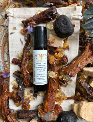 A bottle of Black Cumin Seed, Frankincense and myrrh oil essential oil blend, featuring a black cap and white label, styled on a beige drawstring bag surrounded by dried botanicals, crystals, and a dark stone.