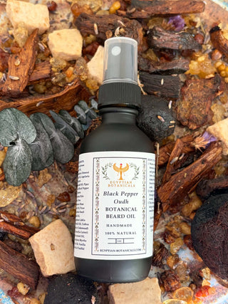 A dark amber glass spray bottle labeled Egyptian Botanicals Black Pepper Oud Botanical Beard Oil' placed on a bed of dried herbs, bark, and stones.