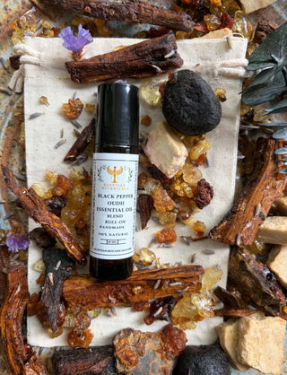 A roll-on essential oil bottle labeled Black Pepper Oudh Natural Botanical Perfume styled in a flat-lay arrangement on a linen cloth surrounded by cinnamon sticks, amber crystals, dried flowers, and a dark stone.