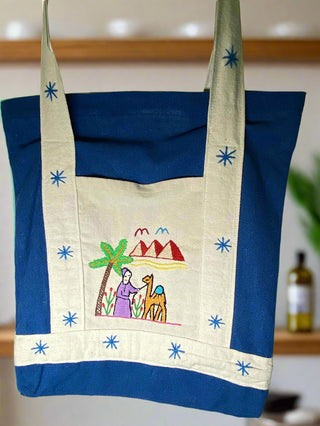 Tote bag with embroidered design of a woman with a camel, palm tree, and mountains.