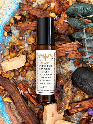 A small amber glass roll-on perfume bottle with a black cap and a cream label reading Egyptian Botanicals Juniper Berry Grapefruit Musk Botanical Perfume, placed on a styled flat-lay of dried herbs, bark, seeds, and a blue textured surface.