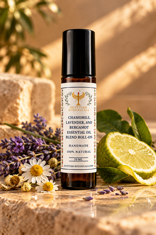 A 10 mL amber glass essential oil roll-on bottle with a black cap and a white label reading 'Egyptian Botanicals' and 'Chamomile, Lavender, and Bergamot Essential Oil Blend Roll-On.' The bottle is styled on a stone surface with chamomile flowers,  bergamot lavender buds, green leaves, and a sliced lime, against a warm beige background.