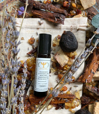 A small black roller bottle labeled Chamomile, Lavender, and Bergamot Natural Botanical Perfume sits on a beige cloth surrounded by dried lavender, cinnamon sticks, amber pieces, and a dark stone. The label reads 'Chamomile Lavender and Bergamot Essential Oil' and '100% Natural Handmade.