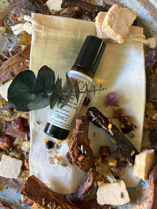 A small amber glass roll-on bottle with a black cap and white label reading 'Create Your Own Essential Oil Blend Roll-On styled on a cream drawstring pouch with dried leaves, bark, seeds, and stone pieces.