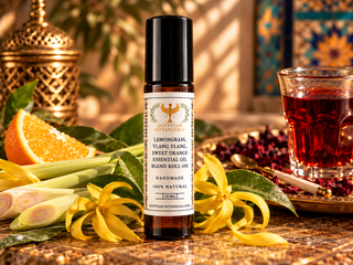 A 10 ml amber roll-on bottle of Lemongrass, Ylang Ylang & Sweet Orange Natural Botanical Perfume surrounded by natural ingredients Lemongrass, Ylang Ylang & Sweet Orange and jojoba oil on a sandy surface beside a glass of tea.

