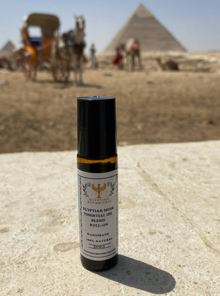 A small roll-on bottle of Egyptian Musk Essential Oil Blend by Egyptian Botanicals stands on a sandy surface with a blurred desert scene and pyramids in the background.