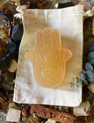 A translucent peach-colored Hamsa Hamsa hand Egyptian Musk Hamsa Soap Bar –Natural Botanical Moisturizing Soap displayed on a small cream fabric drawstring pouch over a rustic background of dried botanicals and stones.