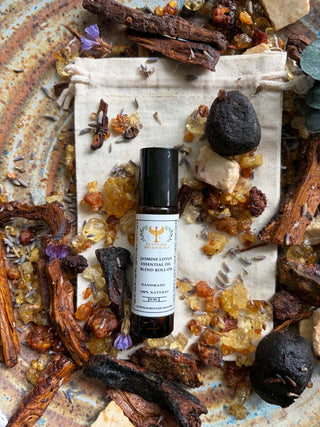A small amber glass roll-on bottle with a black cap and a white label reading 'Jasmine Lotus Natural Botanical Perfume lies on a beige fabric pouch. The pouch is surrounded by dried botanicals, crystals, and dark stones on a rustic metal surface.