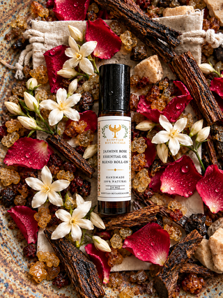 A bottle of Jasmine Rose Essential Oil Blend Roll-On is displayed on a stone ledge surrounded by roses, jasmine flowers, and scattered petals, with a warm, sunlit background.