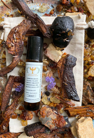A bottle of Lemongrass, Ylang Ylang and Sweet Orange Natural Botanical Perfume roll-on surrounded by dried botanicals, amber crystals, and a small canvas bag on a rustic surface.