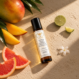 A small amber roller bottle labeled 'Egyptian Mango Grapefruit' sits on a sandy surface surrounded by fruit slices and a white flower. The bottle is the product being sold, while the mango, lime, grapefruit, and flower function as styled props.