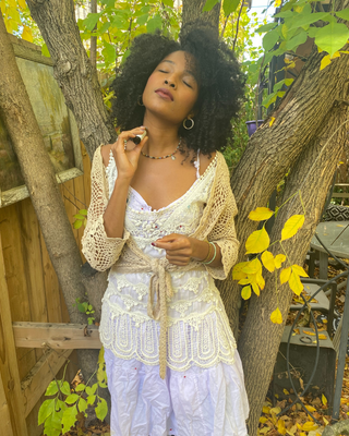 A woman with a large black afro stands outdoors between trees wearing a cream crochet cardigan over a white lace-trimmed camisole and white skirt. She is holding a small black object near her face, with yellow autumn leaves and wooden fencing in the background.