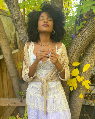 A woman with an afro hairstyle stands outdoors between trees, wearing a cream crochet wrap top over a white lace camisole and light purple skirt. She is appreciating a natural botanical perfume roll on Allspice Oudh Natural Botanical Perfume by Egyptian Botanicals 

