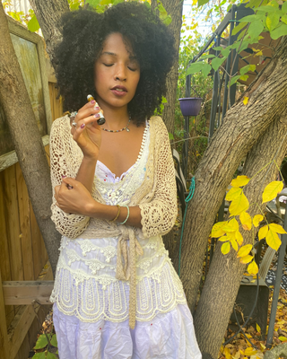 A woman with voluminous dark curly hair stands outdoors in a garden setting, wearing a cream crochet cardigan over a white lace top and a light lavender skirt. appreciating a natural botanical perfume roll on Jasmine Lotus Natural Botanical Perfume by Egyptian Botanicals 

