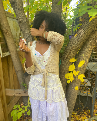 A woman standing outdoors in a garden or backyard setting, wearing a white lace dress with a beige crochet cardigan. She is appreciating a natural botanical perfume roll on Egyptian Mango and Grapefruit Natural Botanical Perfume by Egyptian Botanicals 
