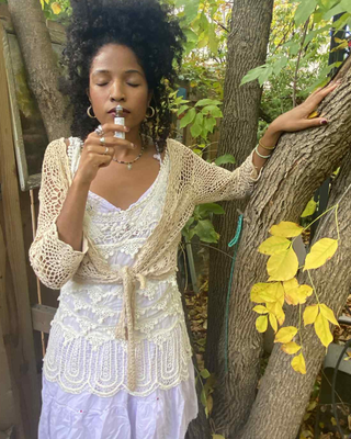 A woman outdoors by a tree wearing a cream crochet cardigan over a white lace top and light purple skirt appreciating a natural botanical perfume roll on Pine Forest-Natural Botanical Perfume by Egyptian Botanicals 
