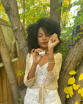 A woman with an afro hairstyle stands outdoors among trees and yellow leaves, holding a small amber glass bottle with a white cap. She wears a white dress and a cream crochet cardigan. She is appreciating a natural botanical perfume roll on Angelica Oudh Natural Botanical Perfume by Egyptian Botanicals 

