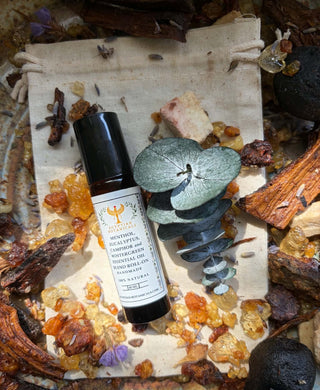 A bottle of Menthol, Eucalyptus, Camphor and Wintergreen Essential Oil Blend Roll-On styled on a beige canvas drawstring pouch surrounded by crystals, dried herbs, and a green eucalyptus leaf.