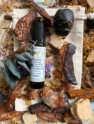 A small roll-on bottle of essential oil surrounded by dried botanicals, crystals, and a small fabric pouch on a rustic stone surface. The bottle label reads Peppermint, Pine and Eucalyptus Essential Oil Blend Roll-On