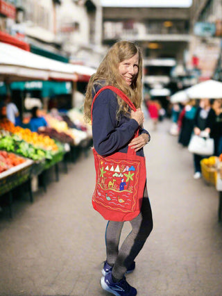 Woman holding a red tote bag with a market scene design, standing in an outdoor market.