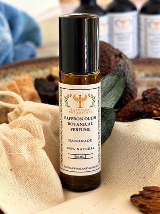 A 10 mL amber glass roll-on bottle of Egyptian Botanicals Saffron Oudh Botanical Perfume with a black cap and white label, styled on a beige linen cloth with dried botanicals and eucalyptus leaves.